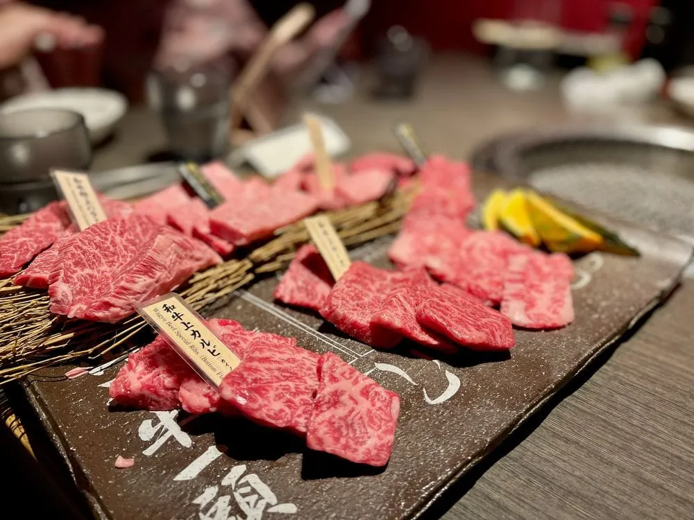 Seared wagyu beef skewers and a grilled wagyu burger patty on a teppanyaki grill, with a water glass and mesh strainer visible on the counter.