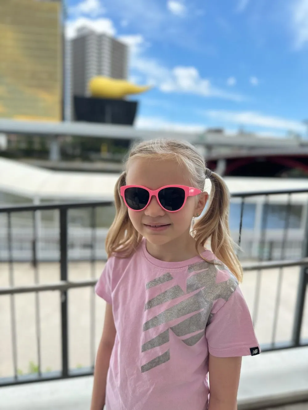 A young girl wearing pink sunglasses stands along the Sumida River in Asakusa, with the golden Flamme d'Or sculpture visible on top of the Asahi building behind her.
