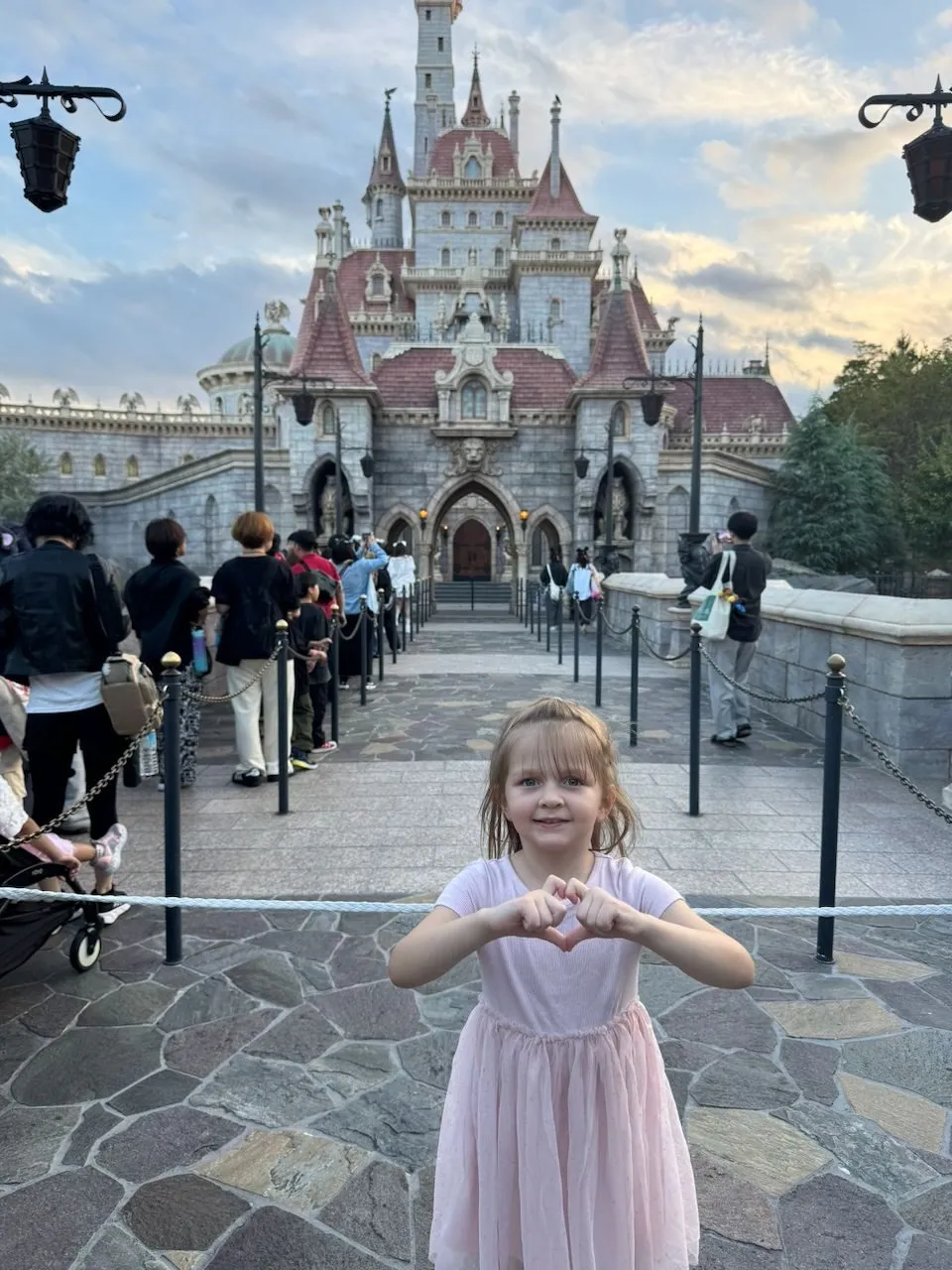 A young girl in a pink dress makes a heart shape with her hands in front of Beast's Castle at Tokyo Disneyland under an overcast sky.