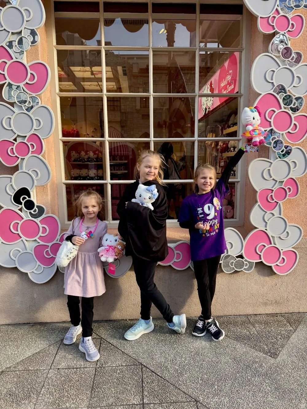Three girls stand in front of the Hello Kitty store at Universal Studios Japan, each holding a Hello Kitty plush, with the oldest wearing a purple Happy 9th Birthday to Me shirt and her arm raised triumphantly.