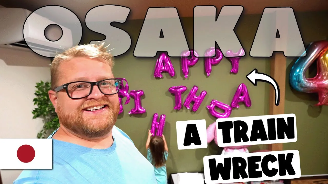 A YouTube thumbnail of Adam Clarkson in an Osaka apartment, a pink foil balloon banner reading jumbled, collapsed letters on the wall behind him, text reading 'A Train Wreck.'