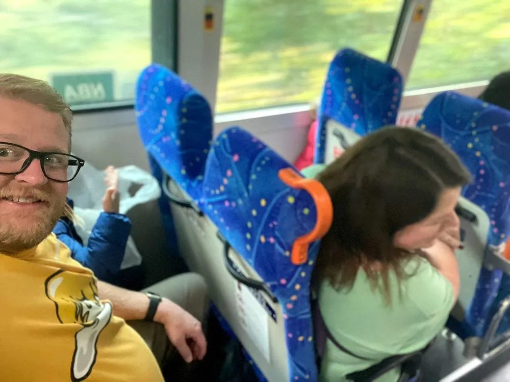 A man in a yellow shirt and glasses grins at the camera from a train seat while a woman beside him laughs with her head down, on a Japanese local train with blue patterned seats and green scenery passing outside the window.