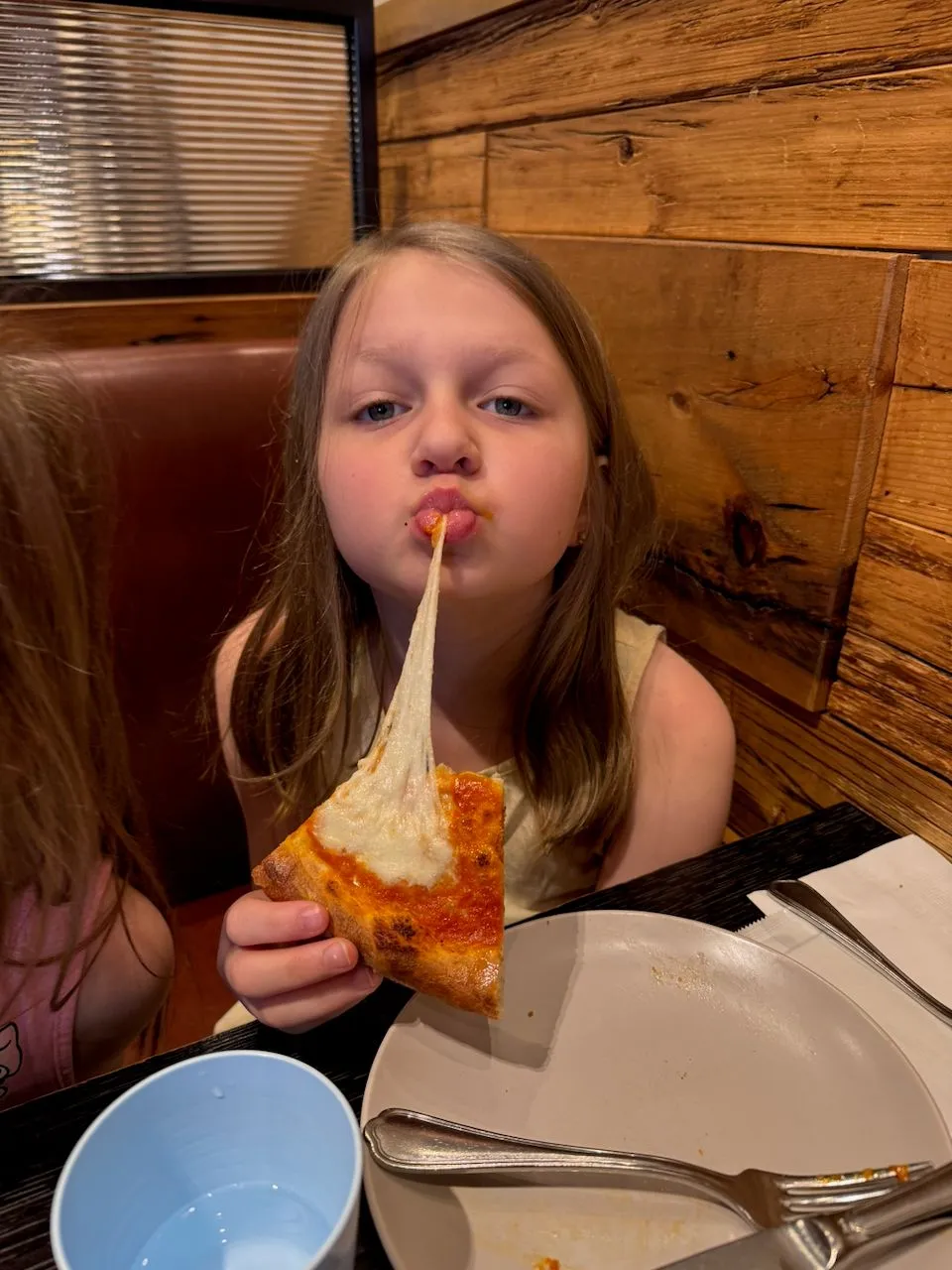 A young girl pulls a long strand of melted mozzarella cheese from a slice of pizza at a casual family restaurant booth in Osaka.
