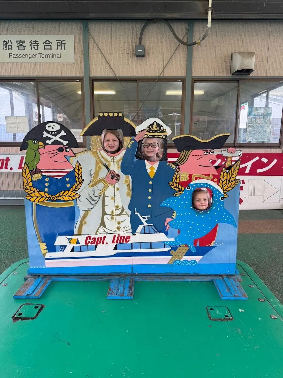 Three children pose with their faces through holes in a painted face-cutout board at a passenger ferry terminal, depicting nautical characters including a ship captain and a dolphin, with Japanese signage visible in the background.