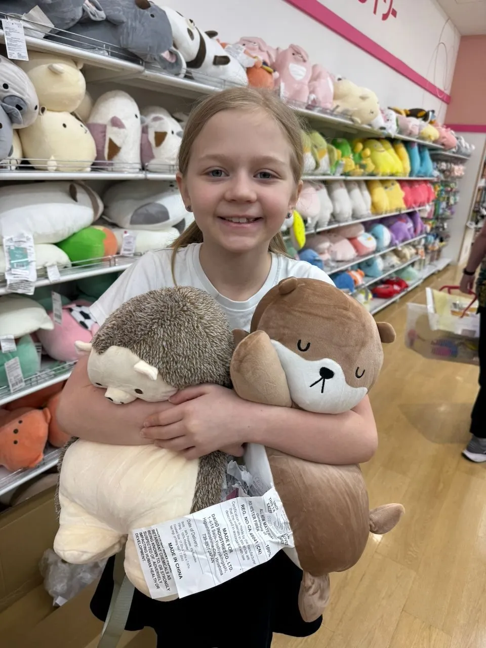 A young girl stands in a colorful Japanese toy shop holding two large stuffed animals, a hedgehog and an otter, while shelves of plush toys line the wall behind her.