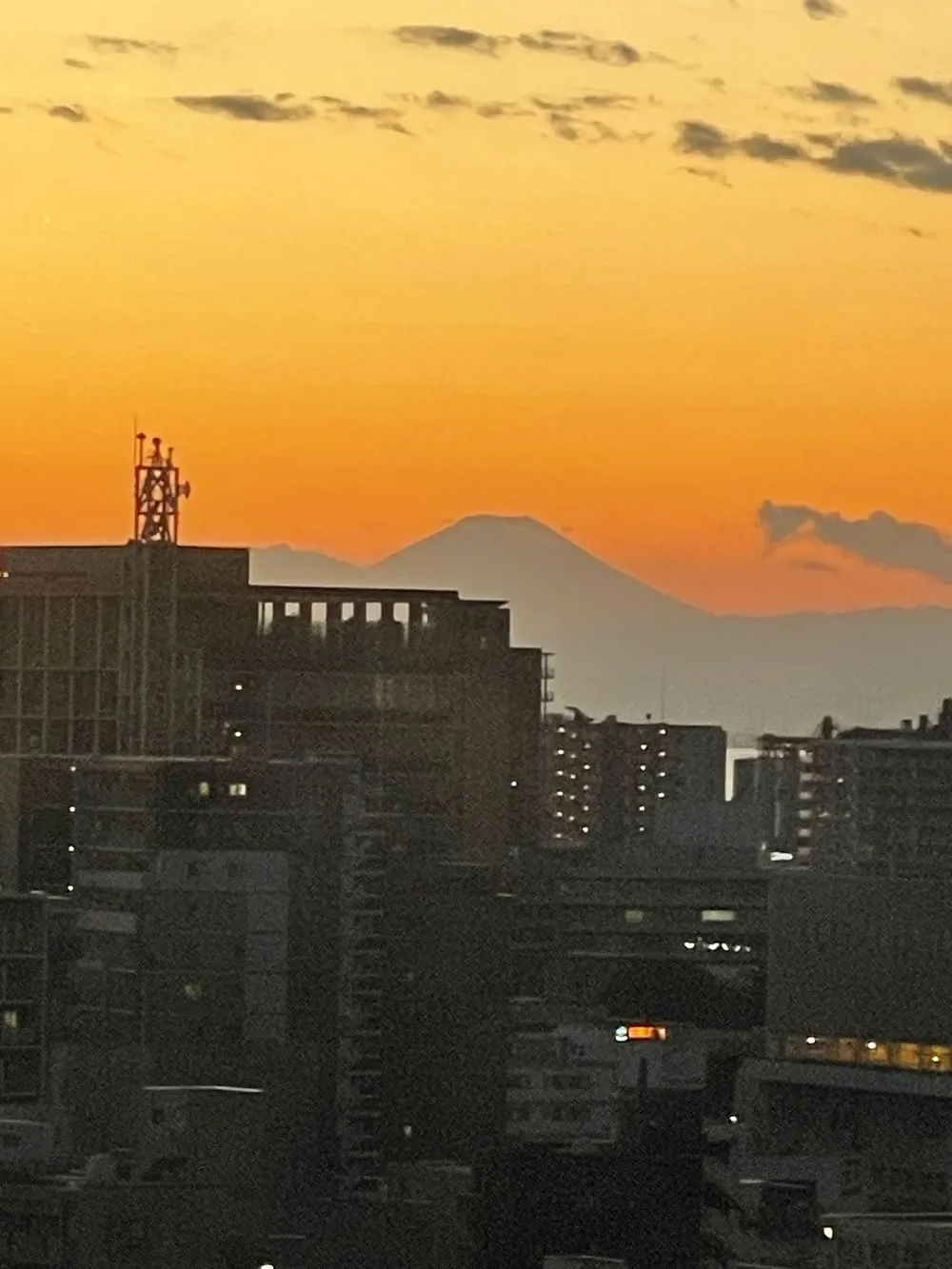 Mount Fuji's silhouette rises above a Tokyo-area skyline at sunset, the sky glowing deep orange behind city buildings.