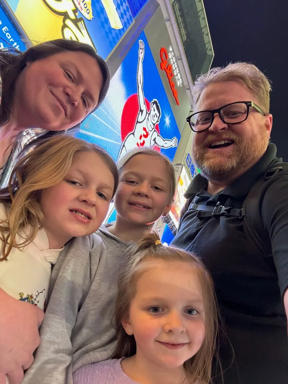 A family selfie of two adults and three young girls in front of the illuminated Glico Man billboard on Dotonbori at night in Osaka, Japan.