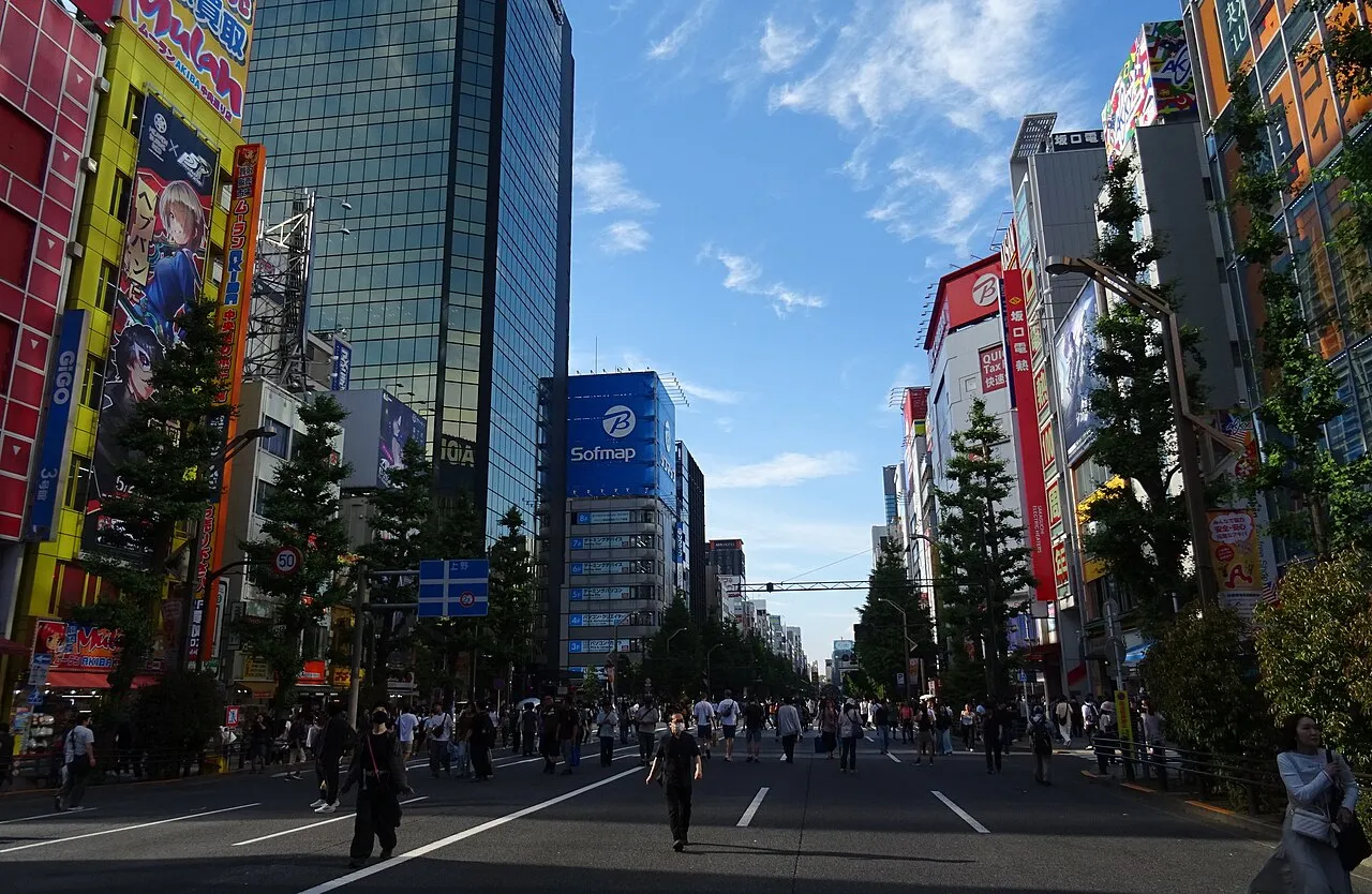 A busy pedestrian street in Akihabara, Tokyo, lined with brightly lit electronics and anime storefronts under a blue sky.