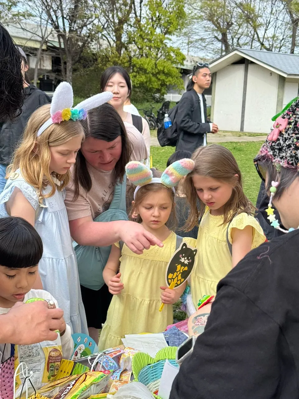 A woman and three young girls wearing bunny ear headbands crowd around an outdoor activity table at an Easter event in Osaka, Japan.