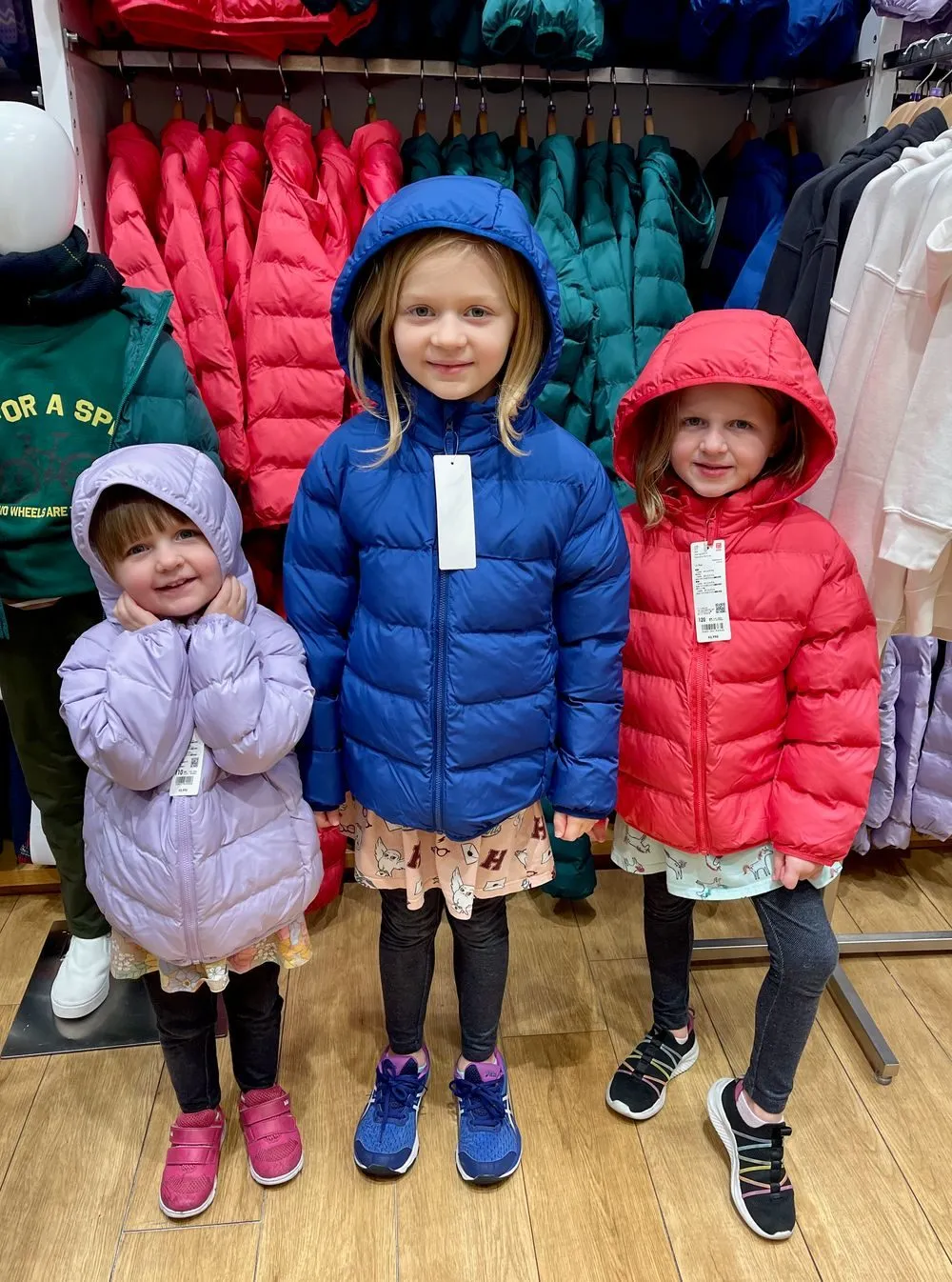 Three young girls try on brightly colored puffer jackets with price tags still attached inside a clothing store, surrounded by racks of winter coats.