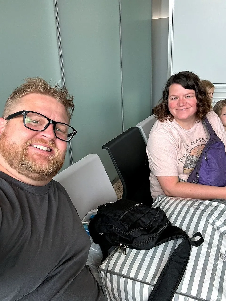 Adam takes a selfie at Narita Airport with Lindsay smiling behind him, bags stacked on the seat between them, a child partially visible beside Lindsay.
