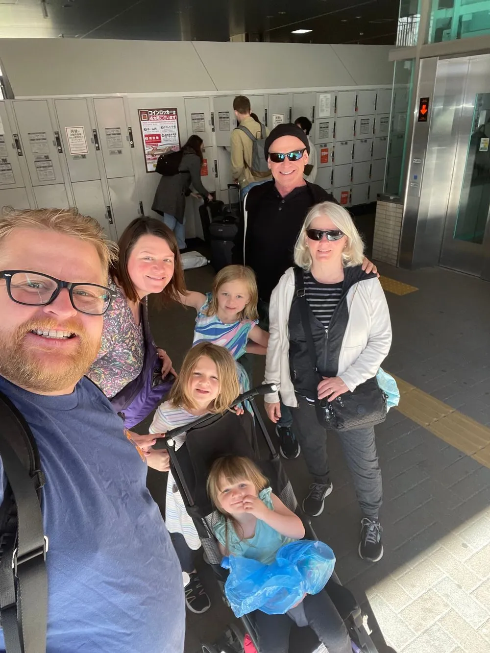 An extended family of seven, including grandparents, poses together in front of coin lockers at a train station, luggage stacked nearby.