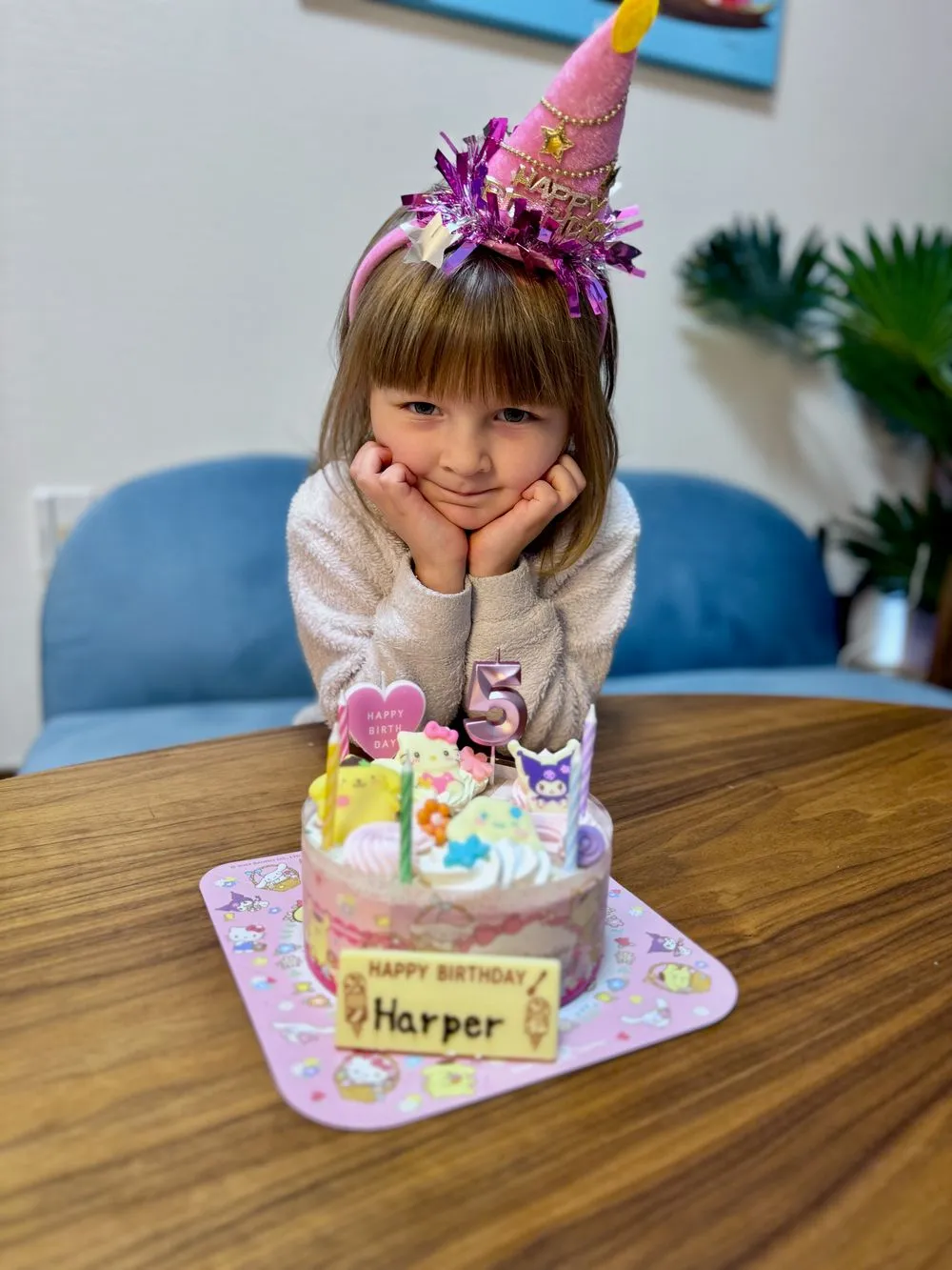 A young girl wearing a pink birthday hat sits with her hands under her chin in front of a decorated Sanrio-themed birthday cake with a nameplate reading Happy Birthday Harper.
