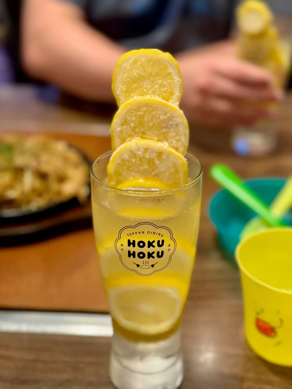 A tall glass of yellow lemon sour drink with four lemon slices stacked on the rim, the glass printed with a HOKU HOKU logo, on a wooden restaurant table.