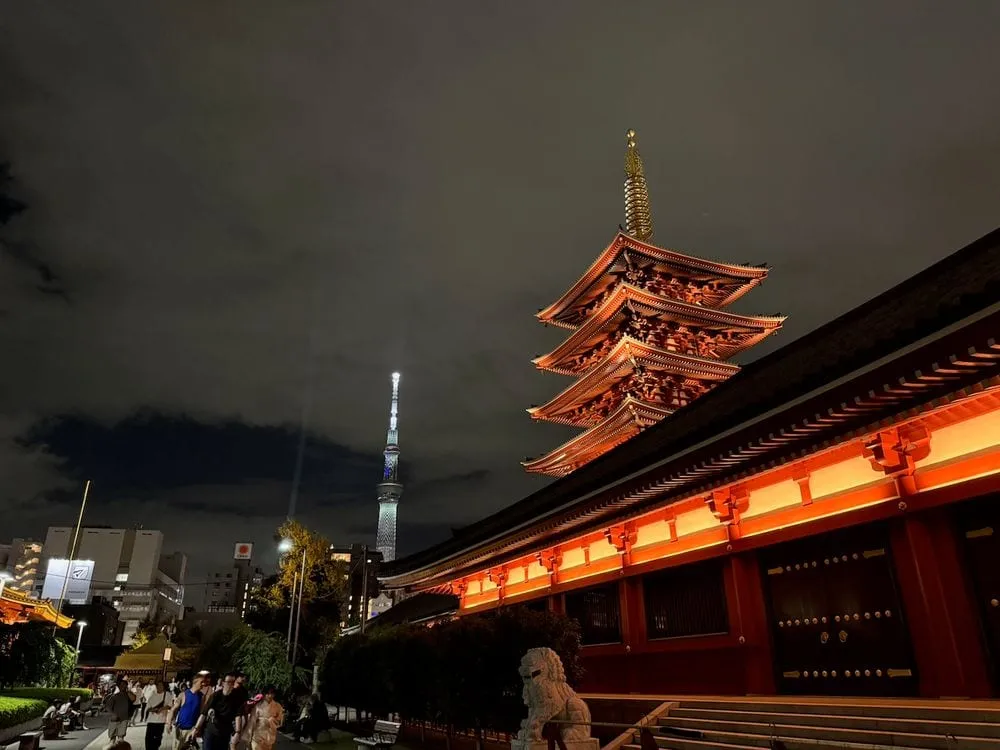 The illuminated five-story pagoda of Senso-ji temple glows orange-red against a dark night sky, with the lit Tokyo Skytree visible in the background.