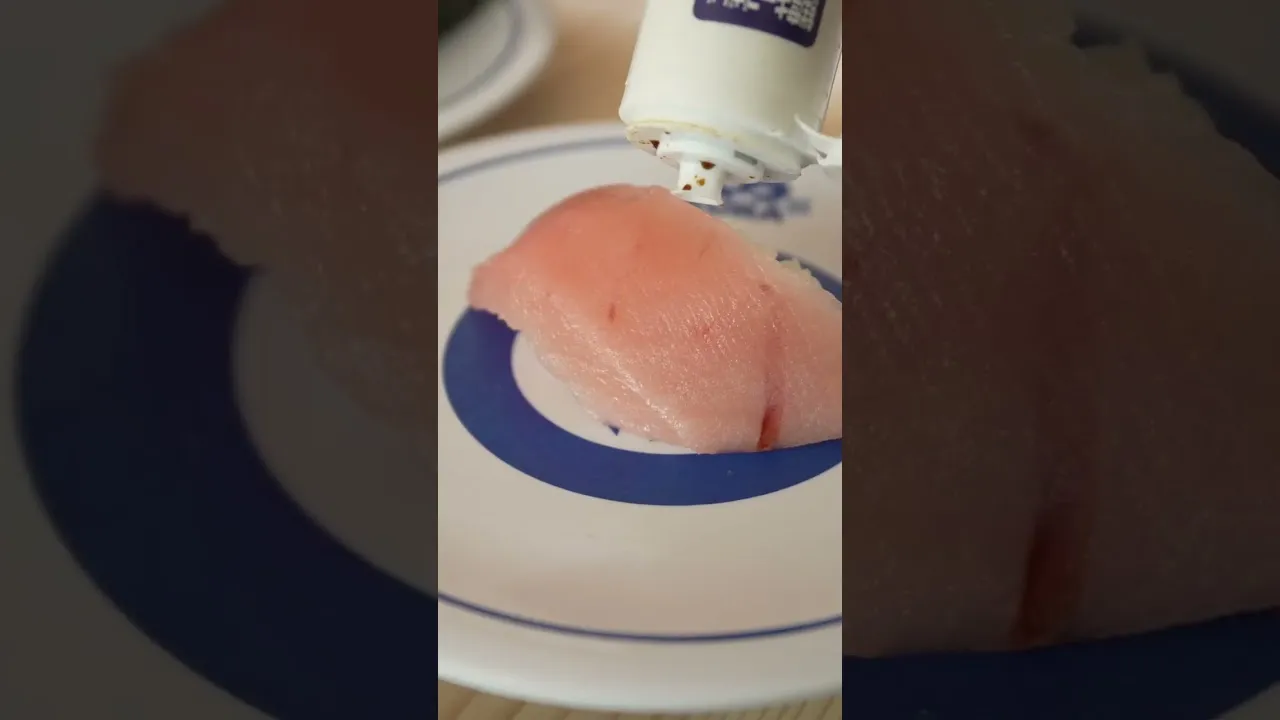 A close-up YouTube thumbnail of a piece of sushi on a blue-patterned conveyor-belt plate at Kura Sushi in Tokyo, with a soy sauce bottle being poured over it.