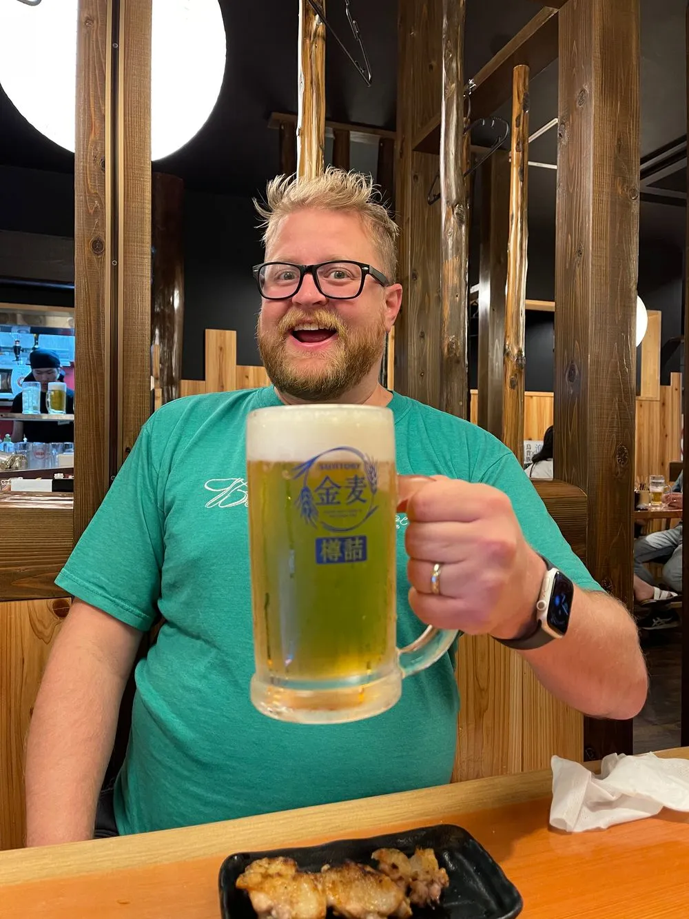 Adam holds up a large frosted mug of draft beer with a grin at an Osaka izakaya, a tray of grilled yakitori skewers visible on the table in front of him.