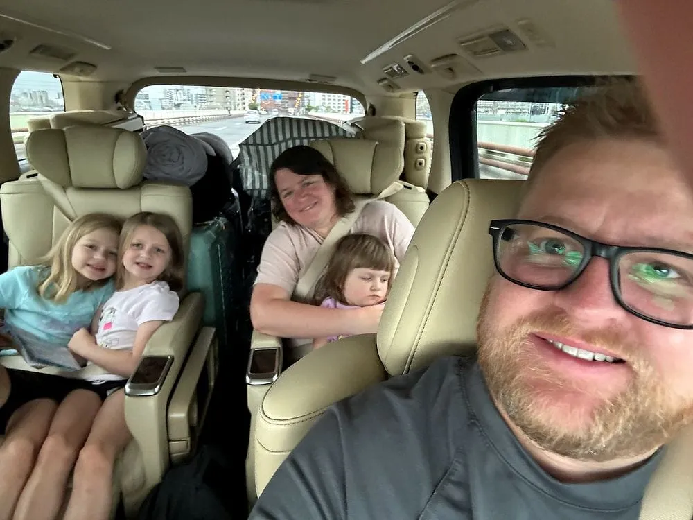 Adam takes a selfie from the front passenger seat of a taxi van, with Lindsay and the three girls visible in the back seats, Harper asleep against Lindsay, and Tokyo visible through the windows.