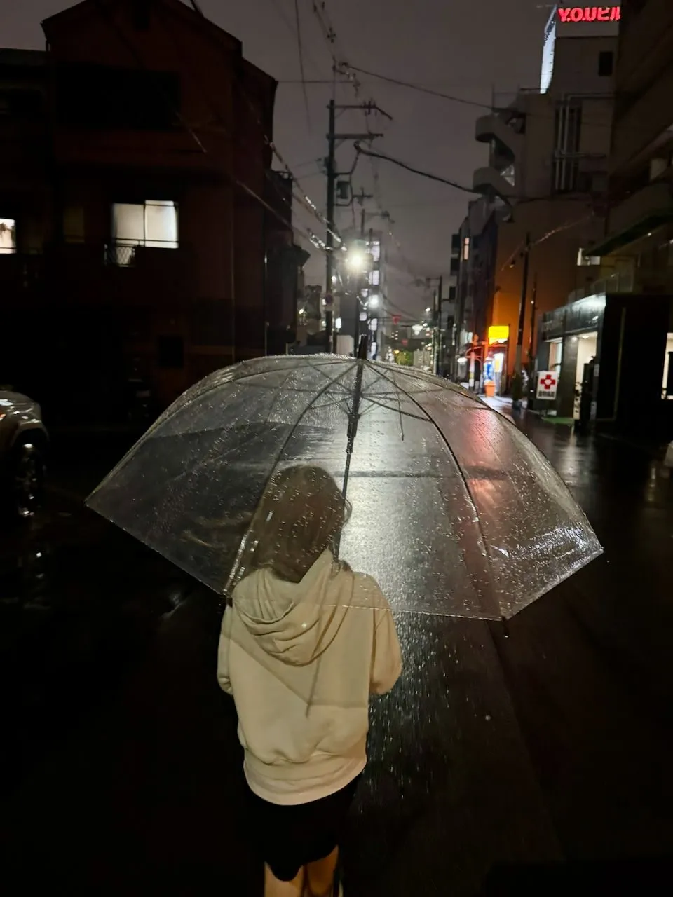 A person seen from behind holding a clear dome umbrella on a rain-slicked Osaka side street at night, with glowing signs visible in the distance.
