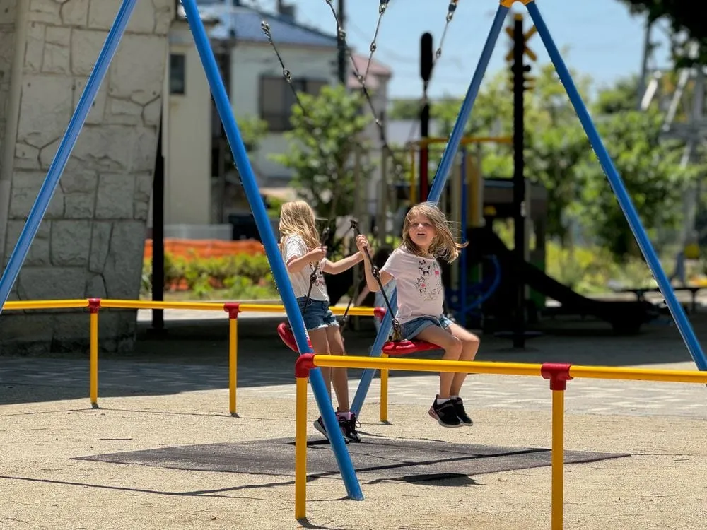 Two young girls play together on a swing set at a neighborhood playground in Katsushika, Tokyo, tucked under an elevated train line, with houses visible in the background.