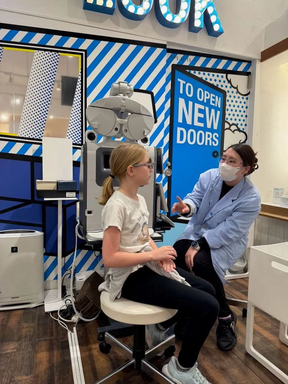 A young blonde girl sits on a stool during an eye exam at a brightly decorated blue and white optician in Osaka, while a masked optometrist gestures and speaks to her beside a phoropter machine.