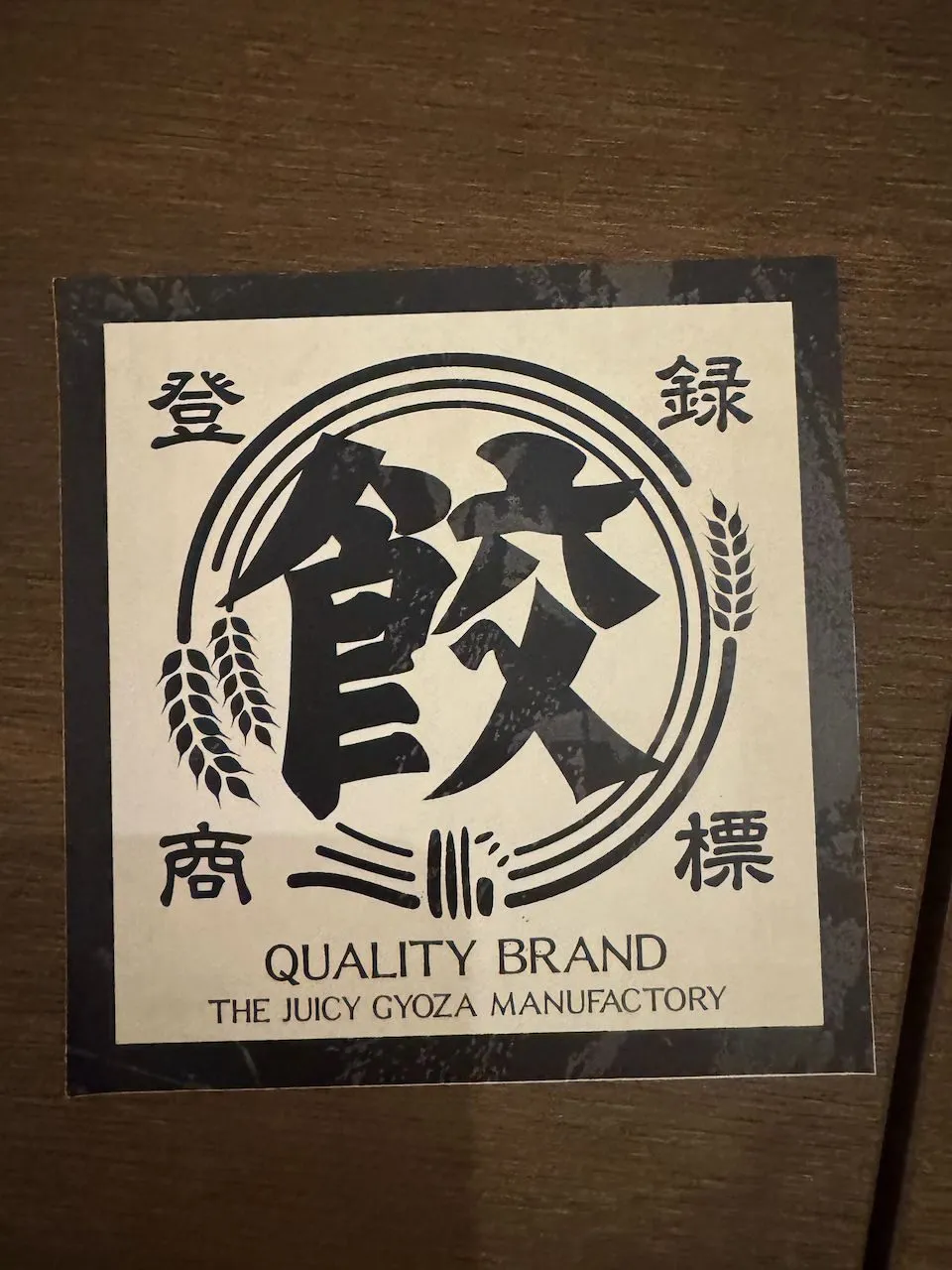 A square paper coaster on a wooden table, printed with bold Japanese kanji and the English text 'Quality Brand, The Juicy Gyoza Manufactory.'