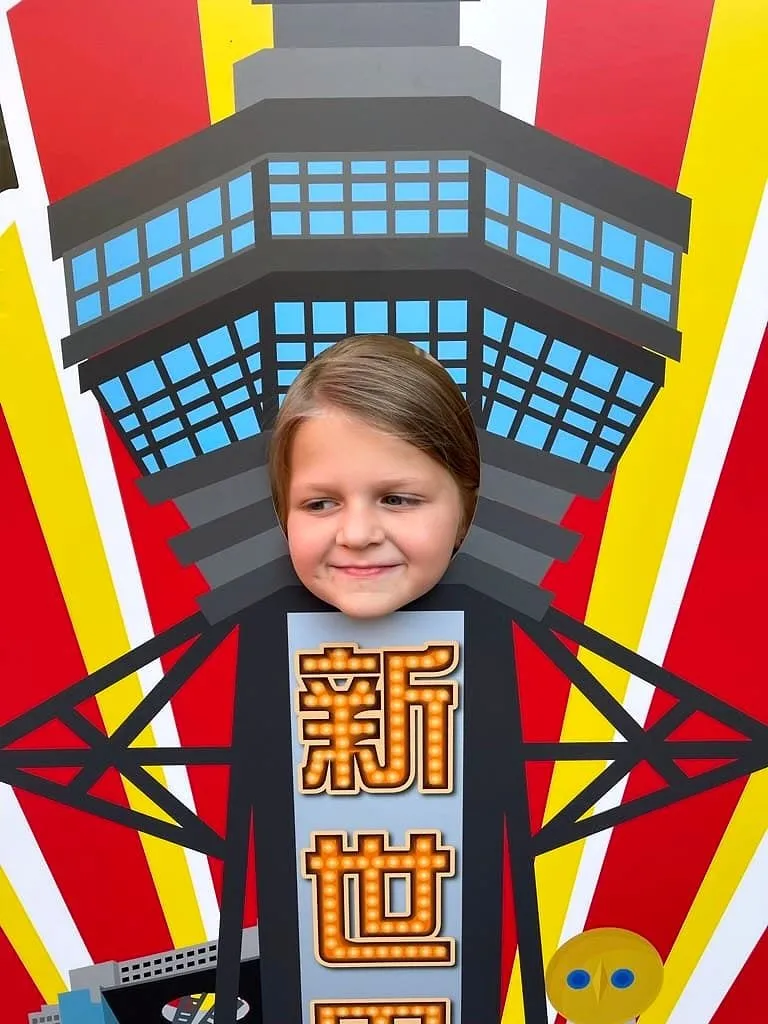 Cora's face peeking through a face cutout board shaped like Tsutenkaku Tower on a red and yellow illustrated sign in Shinsekai, Osaka.
