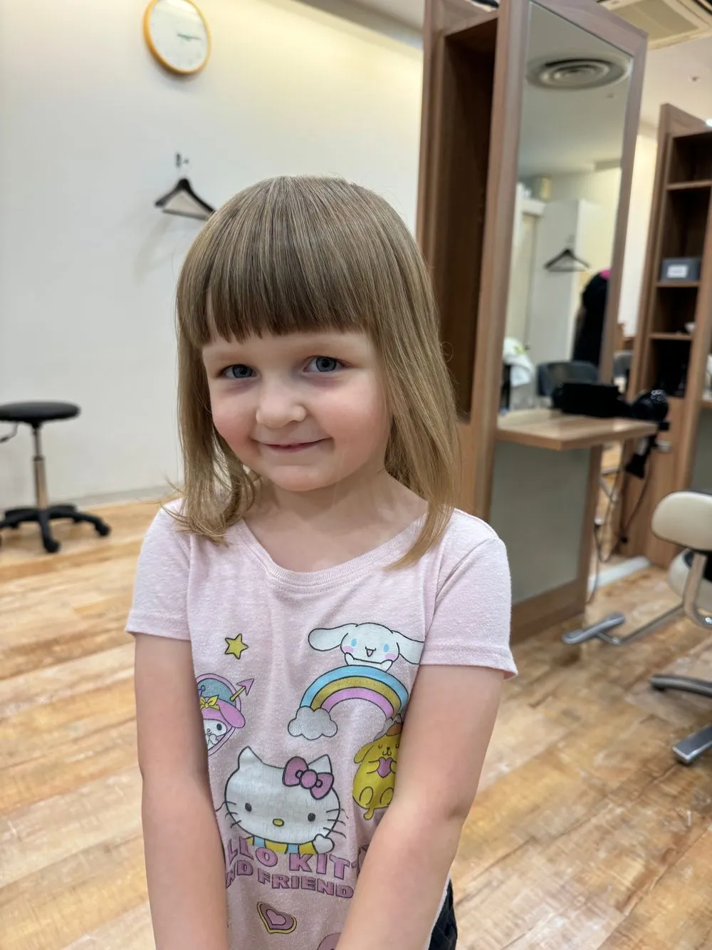 A young girl with fresh-trimmed bangs smiles in a Japanese hair salon, wearing a Hello Kitty shirt, with salon chairs and a mirror visible behind her.