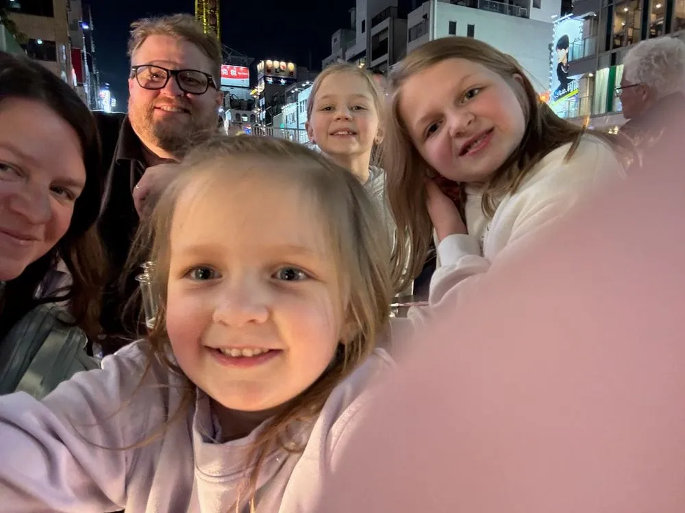 A family selfie at night on a neon-lit street in Osaka's Sōemon district, with two parents and three young girls smiling at the camera and illuminated signs visible in the background.
