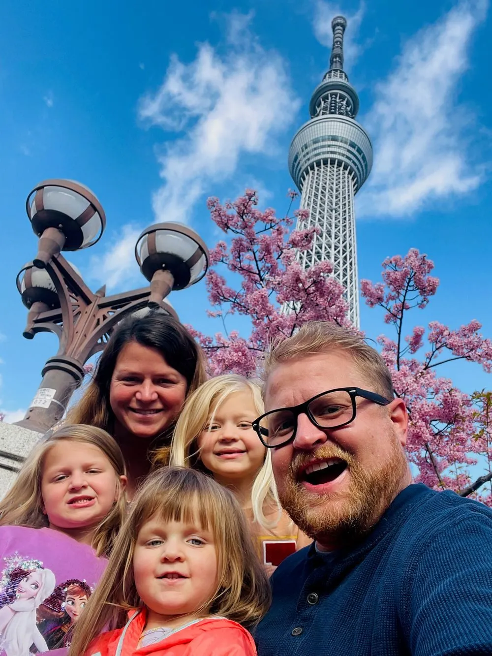 A family selfie of five people in front of Tokyo Skytree, with pink cherry blossoms blooming behind them against a blue sky.