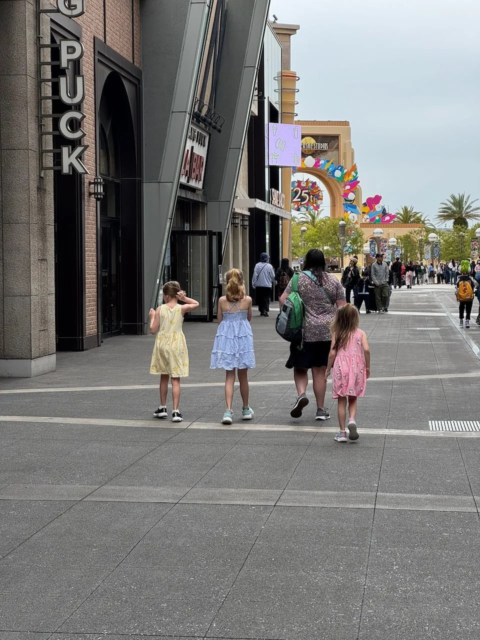Lindsay and three young girls in summer dresses walk toward the Universal Studios Japan entrance arch, with colorful 25th anniversary decorations visible ahead.