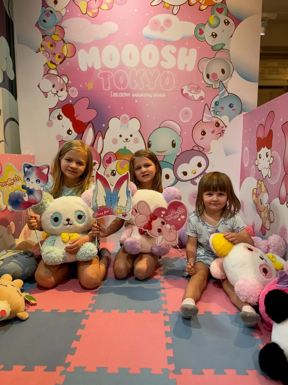 Three young girls kneel together holding large squishy plush toys inside MOOOSH TOKYO, a pink character-themed store in Harajuku, surrounded by shelves of squishy characters.