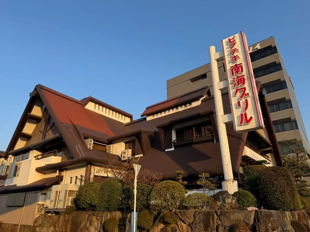 A low-angle exterior shot of a large Japanese restaurant building with a steep dark-roofed alpine-style facade and a tall vertical sign in red kanji reading Nankai Grill, shot in warm late-afternoon light against a clear blue sky.
