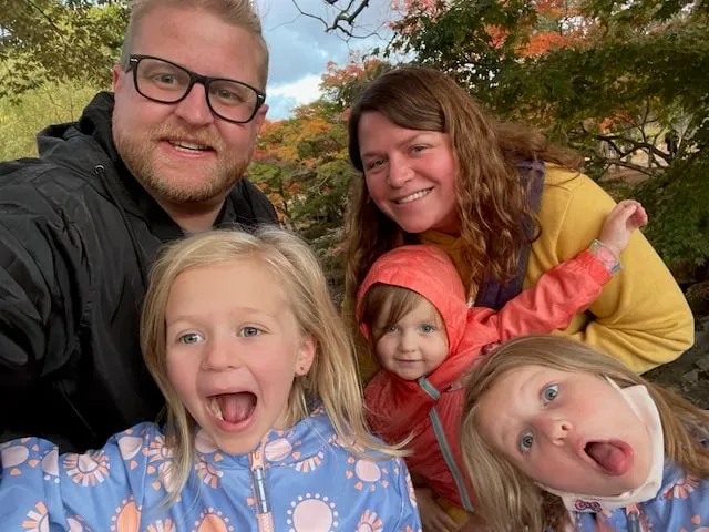 A family of five takes a selfie in Nara, Japan, with autumn foliage visible behind them, two of the girls making silly faces at the camera while the parents smile.