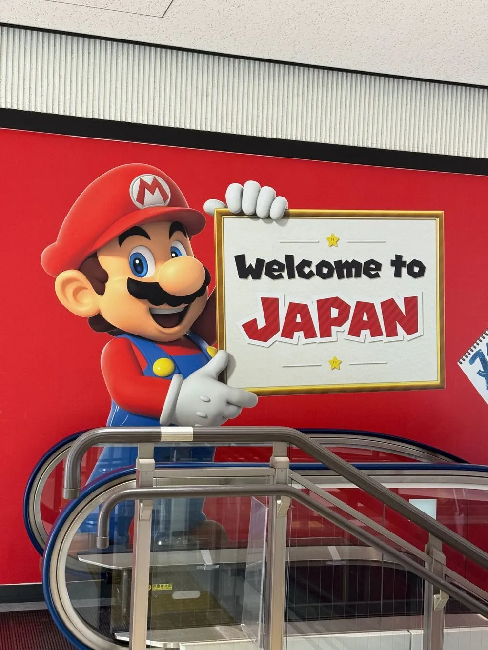 A large Mario character display stands beside an escalator at Narita Airport, holding a sign that reads 'Welcome to Japan.'