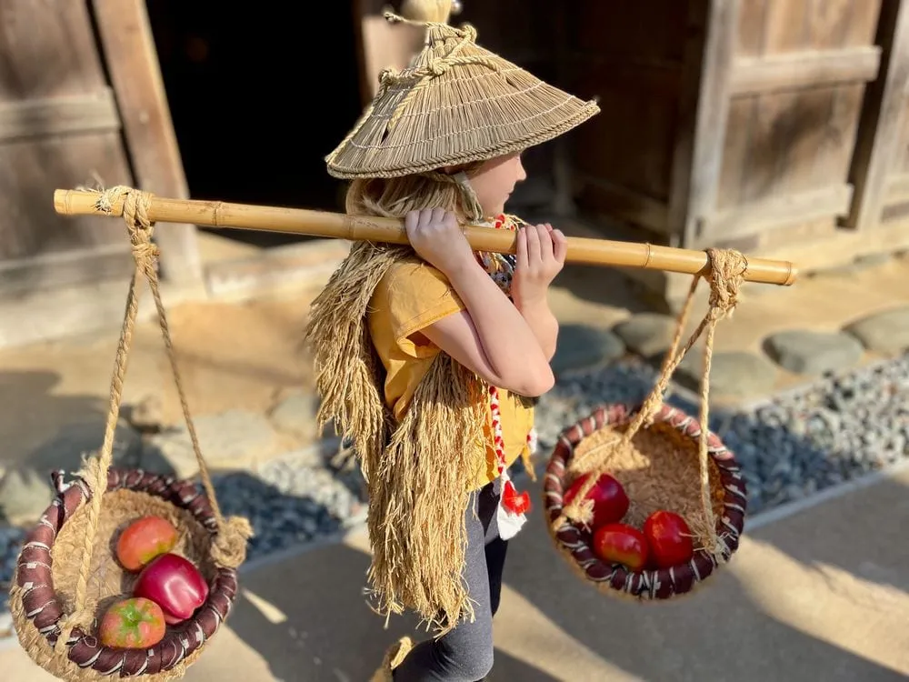 A young girl in a straw hat and cape carries a wooden yoke with two baskets across her shoulders in front of a historic thatched-roof farmhouse at Nihon Minkaen in Kawasaki.
