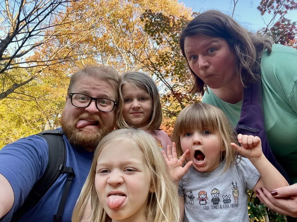 Adam takes a selfie with Lindsay and their three daughters making silly faces, surrounded by orange and yellow autumn foliage at Nihon Minkaen in Kawasaki, Japan.