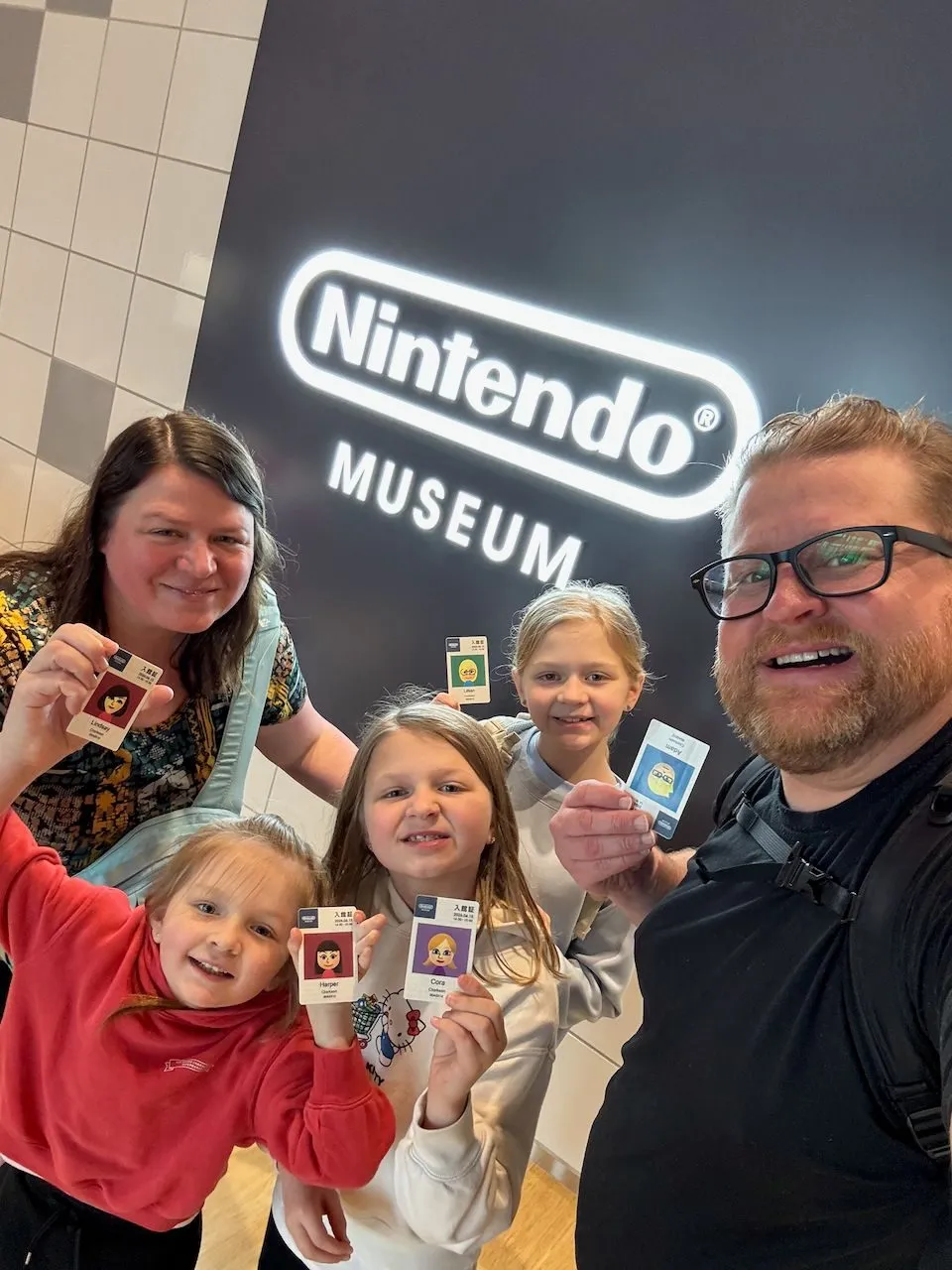 A family of five poses in front of the illuminated Nintendo Museum sign, each holding up their personalized character entry cards.