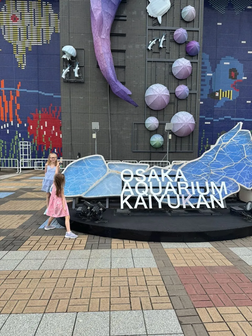 Two young girls stand in front of the large Osaka Aquarium Kaiyukan entrance sign, decorated with a whale shark sculpture and ocean-themed mosaic tiles, one waving at the camera.