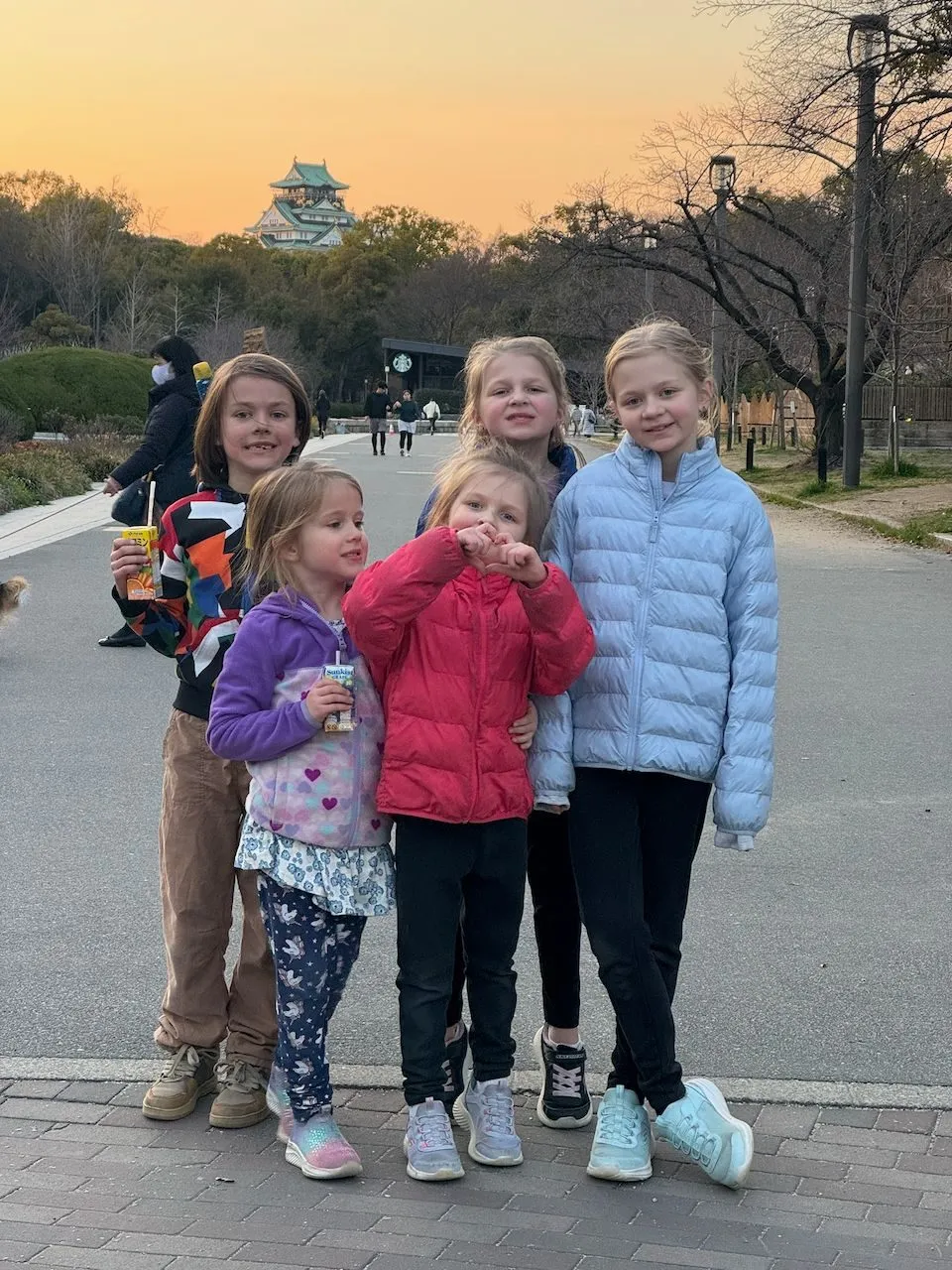Five girls pose together on a path in Osaka Castle park at sunset, with the castle visible in the warm orange sky behind them.