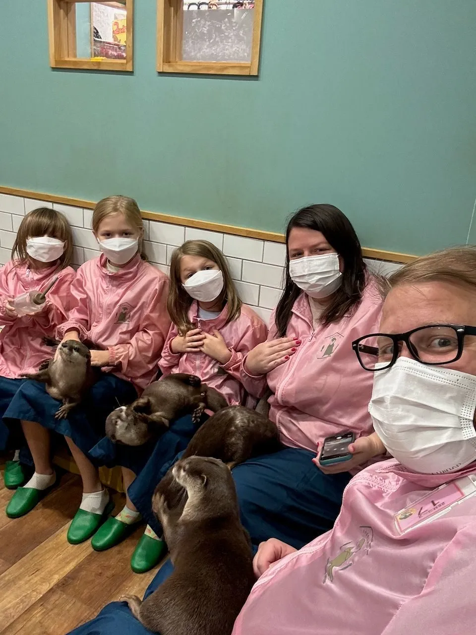 Lindsay and three girls sit in a row wearing matching pink cafe jackets and face masks at an otter cafe in Harajuku, each with a small brown otter on or near their lap, while a fifth otter reaches up toward the camera in the foreground.