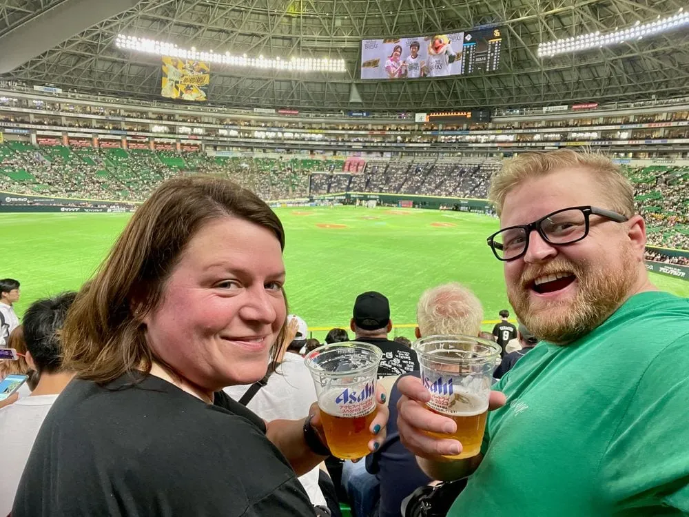 Adam and Lindsay smile at the camera holding cups of Asahi beer in the stands of a packed domed baseball stadium in Fukuoka, with the bright green field visible behind them.