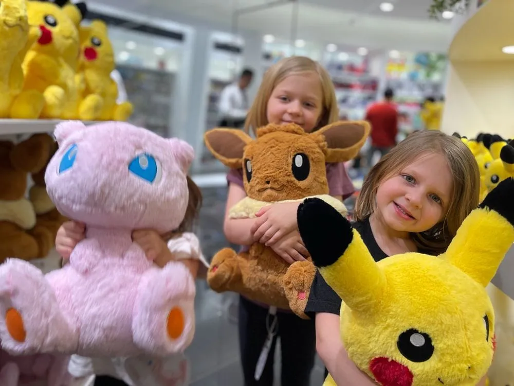 Two young girls hold up stuffed Pokemon plush toys, including a pink Mew, an Eevee, and a yellow Pikachu, inside a brightly lit store.