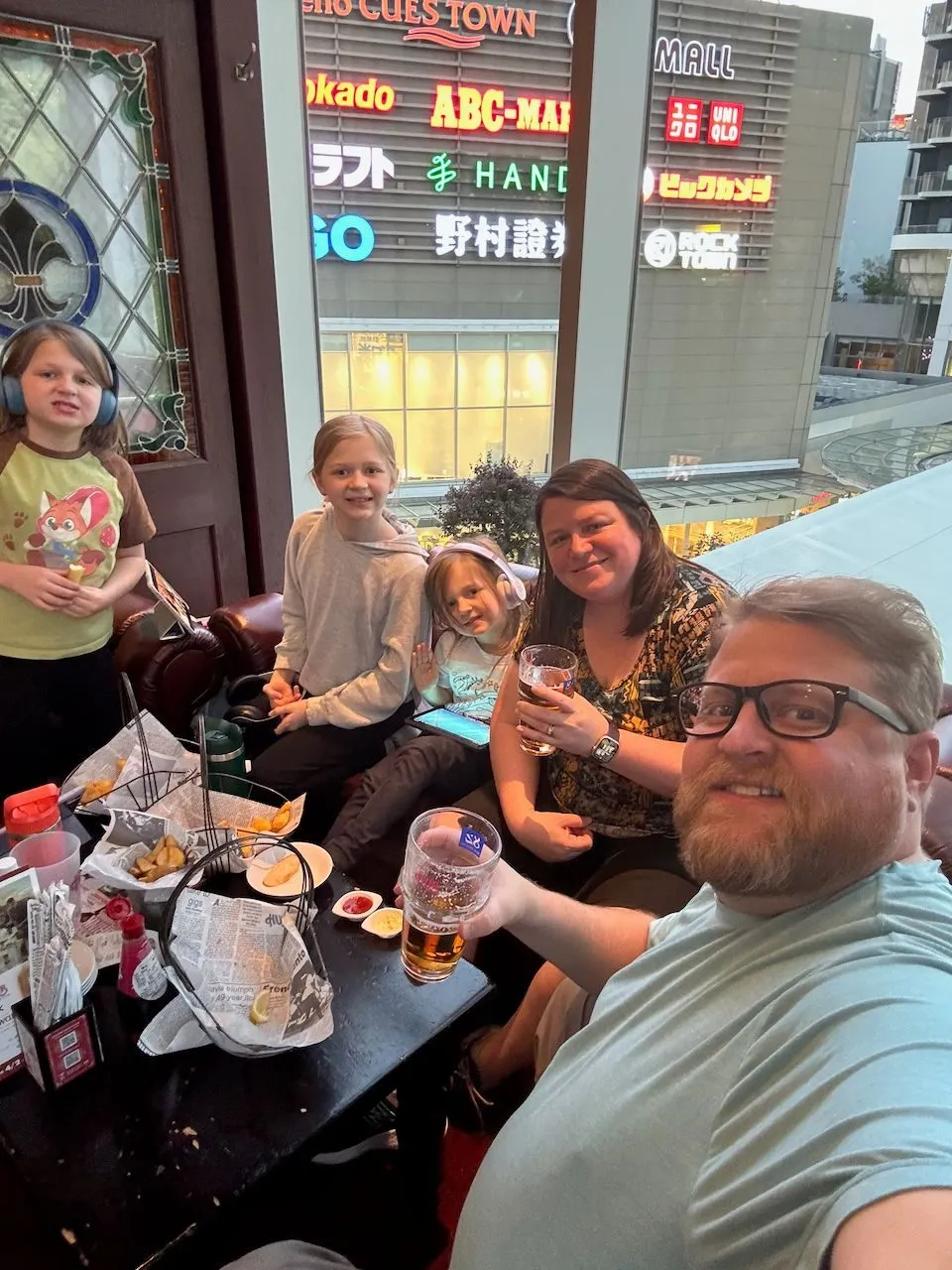 Adam takes a selfie at a pub table in Osaka with Lindsay holding a beer beside him, Lily and Harper seated across the table, and Cora standing to the left wearing headphones, with neon shopping signs visible through the window behind them.