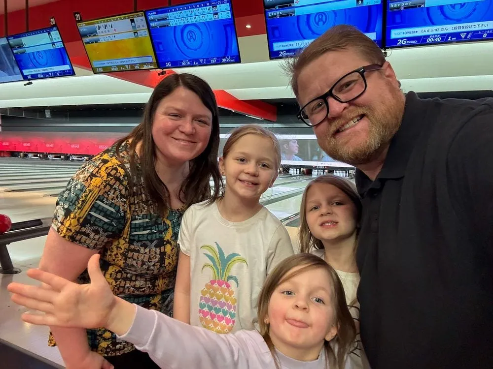 A family of five poses for a selfie at a Japanese bowling alley, with lane scoreboards visible overhead showing names in both English and Japanese.