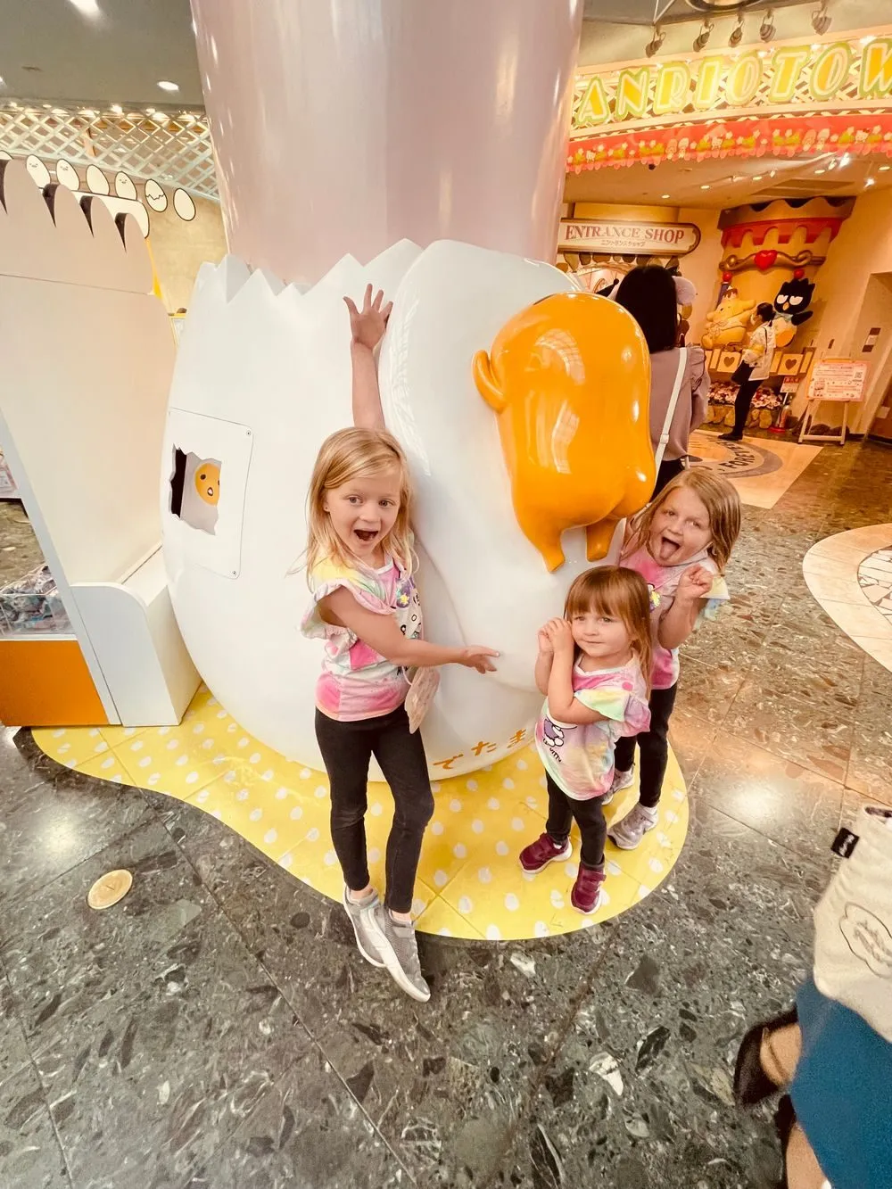 Three young girls pose and make silly faces next to a large Gudetama egg sculpture inside Sanrio Puroland.
