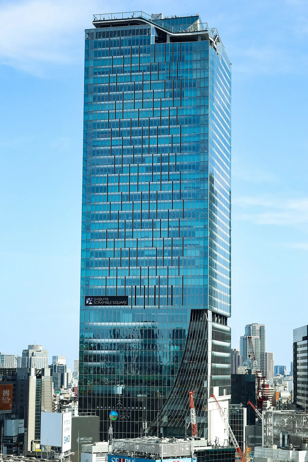 The glass tower of Shibuya Scramble Square in Tokyo, home to the Shibuya Sky observation deck, seen from street level under a clear blue sky.