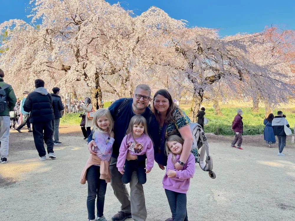 A family of five, two adults and three young girls, pose smiling in front of large weeping cherry blossom trees in full bloom at a crowded park on a sunny day.