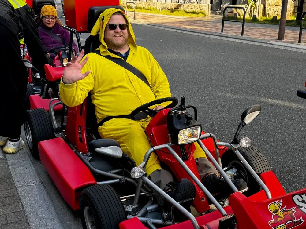 Adam, dressed as Pikachu, waves from the driver's seat of a red street kart stopped on a road in Asakusa, Tokyo.