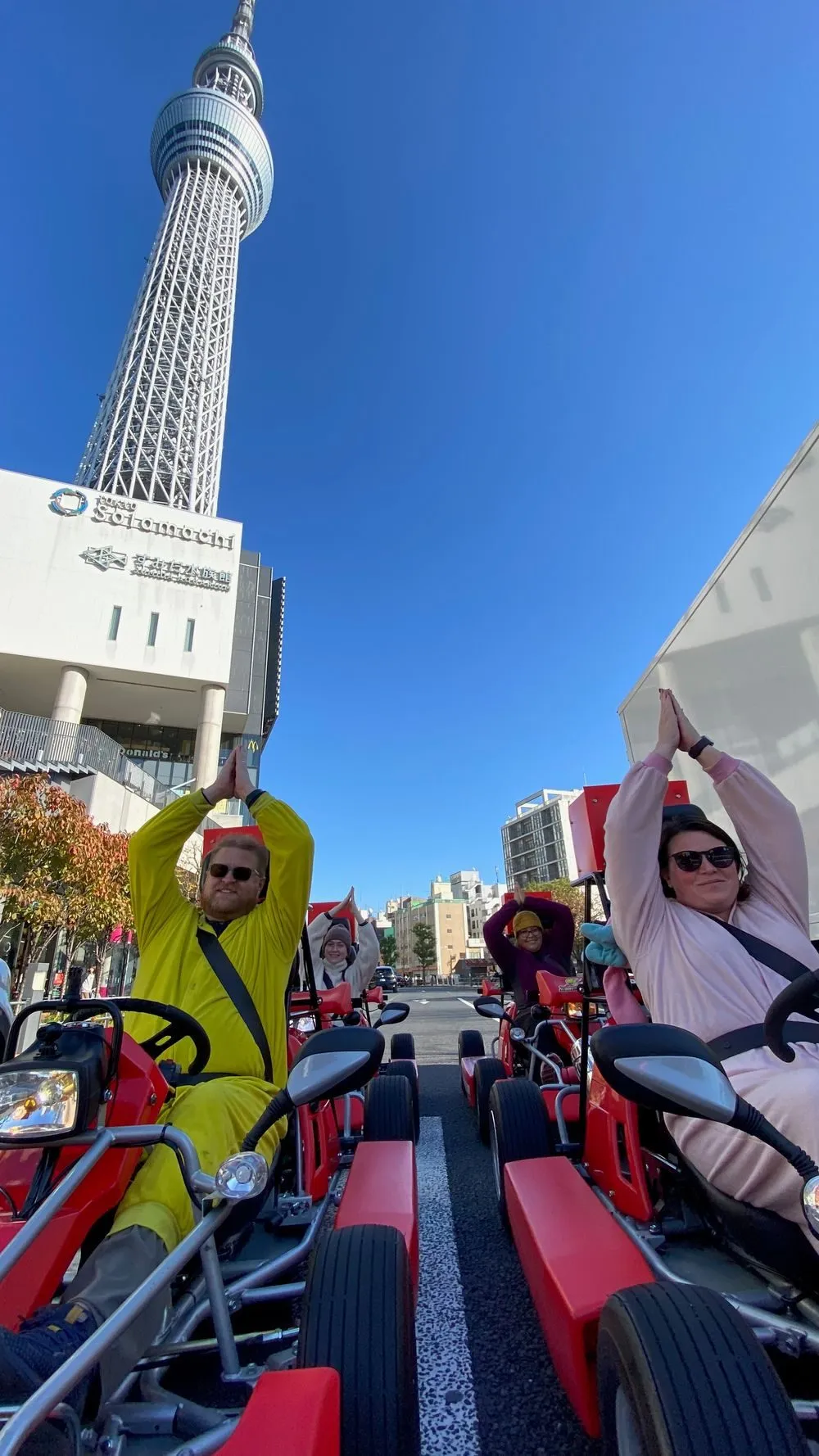 Adam in a yellow Pikachu costume and Lindsay in a pink My Melody costume sit in red street karts with arms raised overhead, Tokyo Skytree towering directly behind them against a clear blue sky.